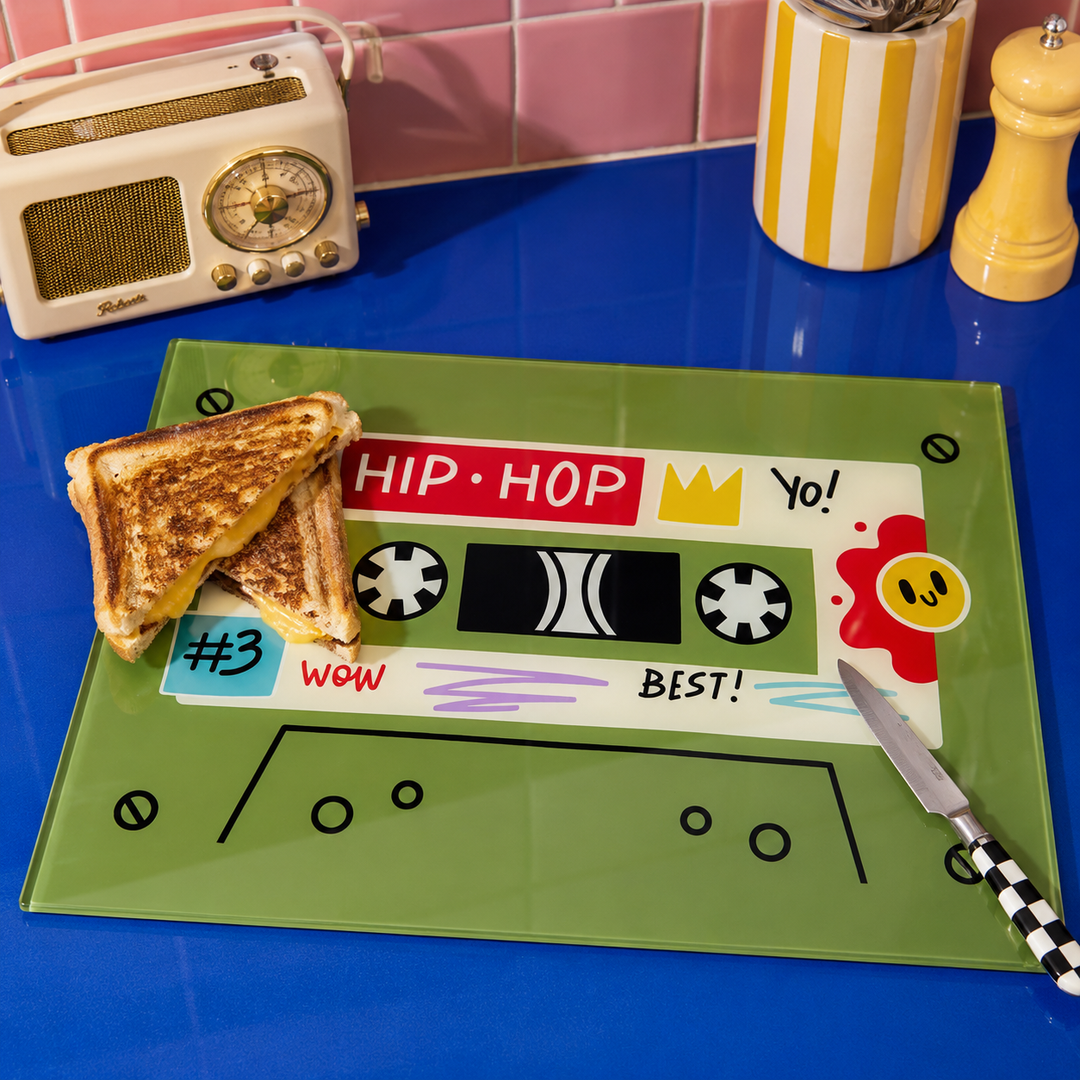 A green glass cutting board with a retro cassette tape design, styled on a blue countertop with a knife, grilled cheese, and a vintage radio in the background.