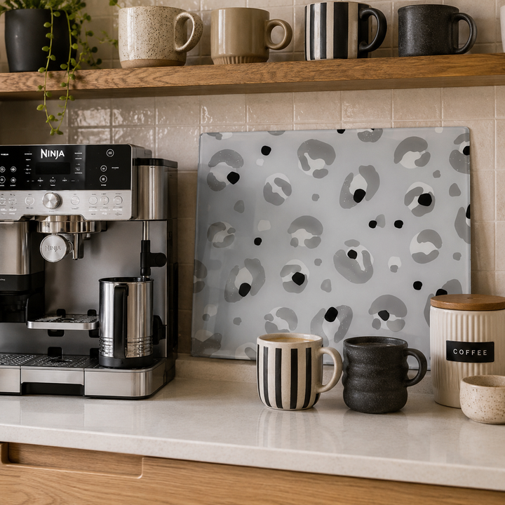 A rectangular glass chopping board with a gray, black, and white organic spot pattern leans against a tiled kitchen wall next to a coffee machine and ceramic mugs.