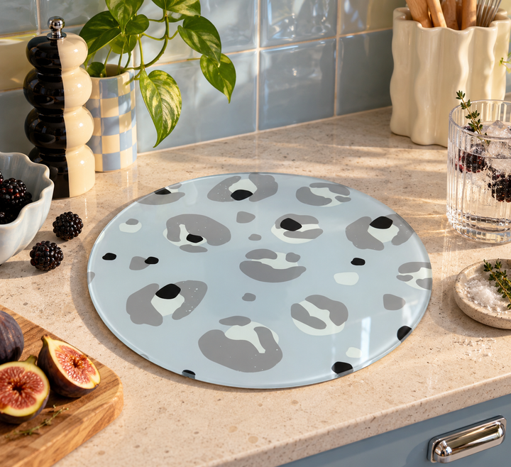 A round light grey glass worktop saver with a gray, white, and black organic spot pattern, styled on a beige kitchen countertop with fresh figs, a plant, and kitchen accessories.