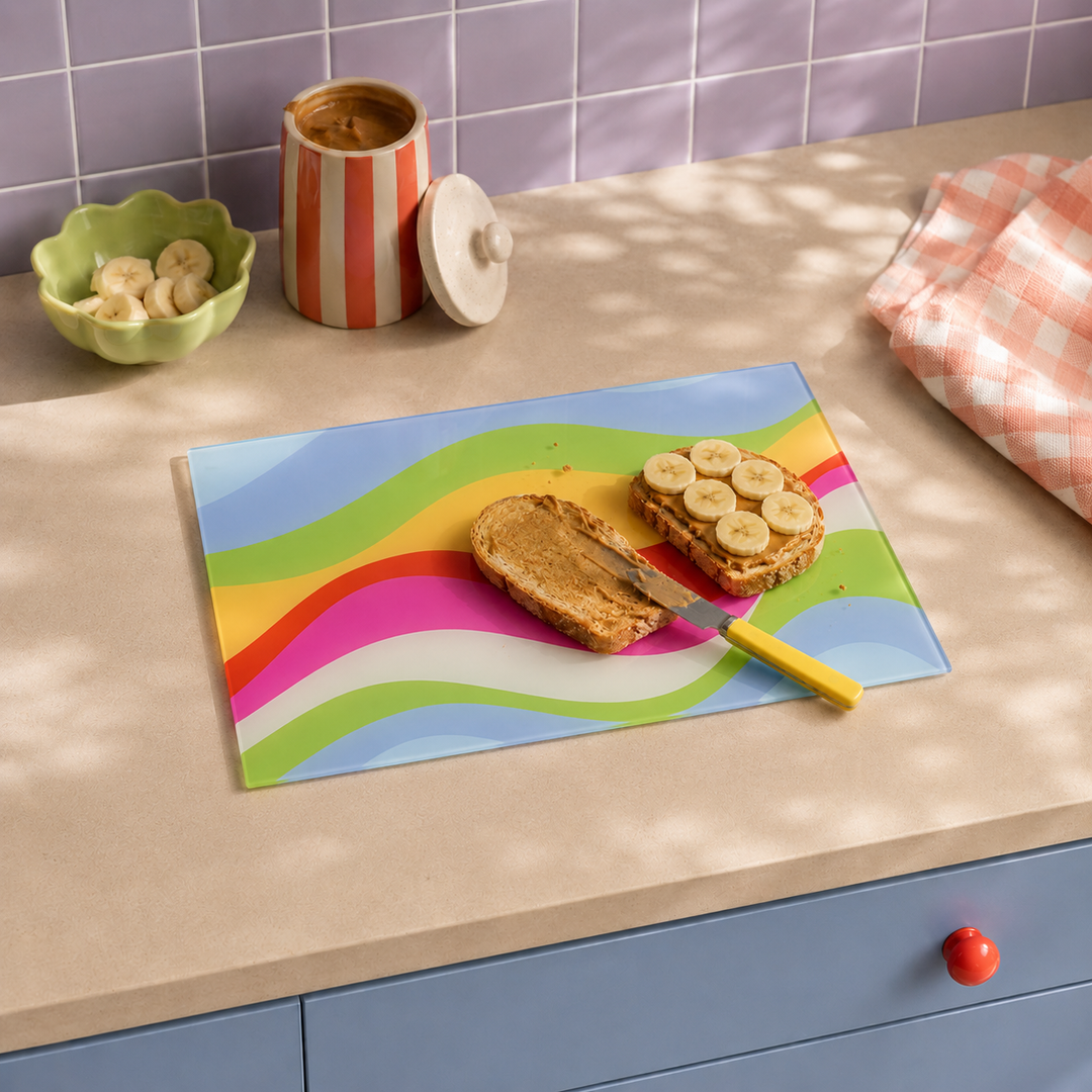 A colorful wavy-patterned cutting board on a beige kitchen countertop, with a slice of toast and banana-topped toast on it and a yellow-handled knife resting on the board. The scene includes a green bowl of banana slices, a striped jar, and a pink-and-white checkered cloth, with purple tiled backsplash and blue cabinets in the background.