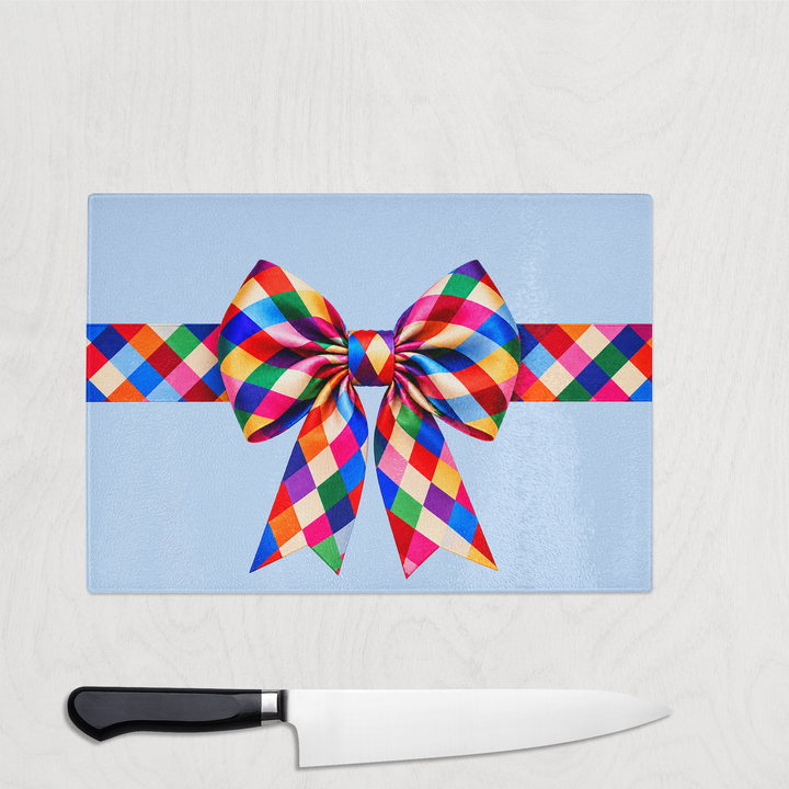 Harlequin Colourful Bow Christmas glass chopping board with a large rainbow harlequin check ribbon and bow on a pale blue background, shown with a chef's knife