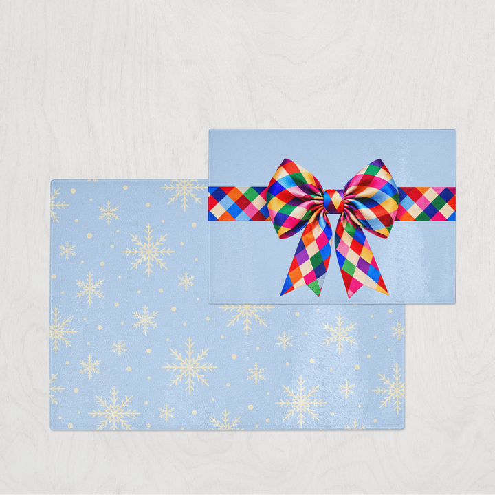 A light blue gift box style glass chopping board with white snowflake patterns, wrapped with a vibrant multicolor plaid ribbon tied in a bow, set against a light gray wood-textured background.