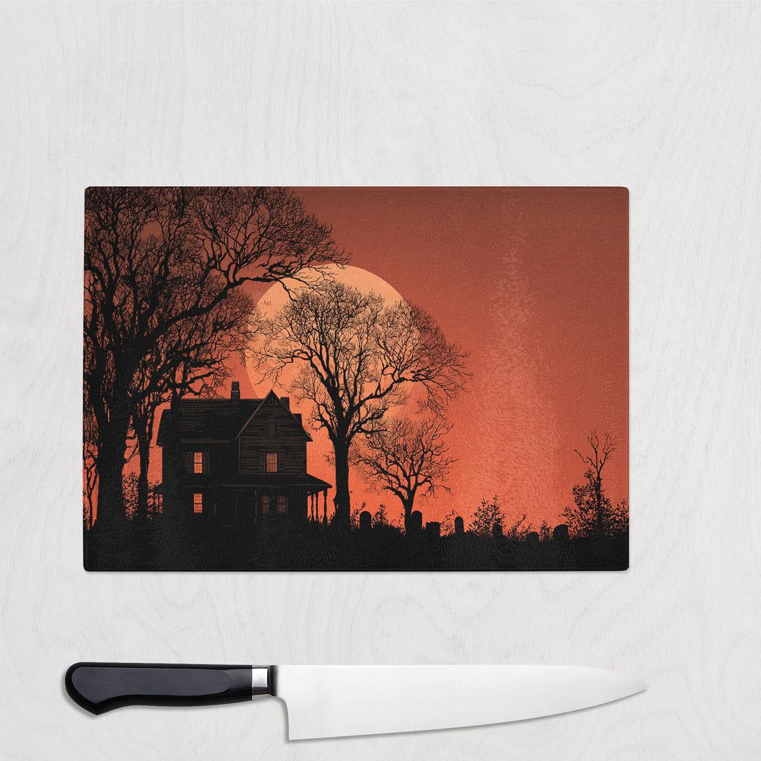 Haunted House At Dusk glass chopping board with a silhouetted old house, bare trees and headstones against a burning orange sky