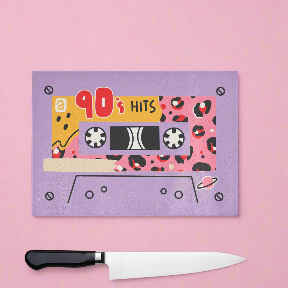 The Yililo Lilac 90s Hits Cassette Tape Chopping Board adds retro charm to your kitchen with bold patterns and 90s Hits text. Perfect for nostalgic 90s fans, it rests on a pink surface next to a kitchen knife.