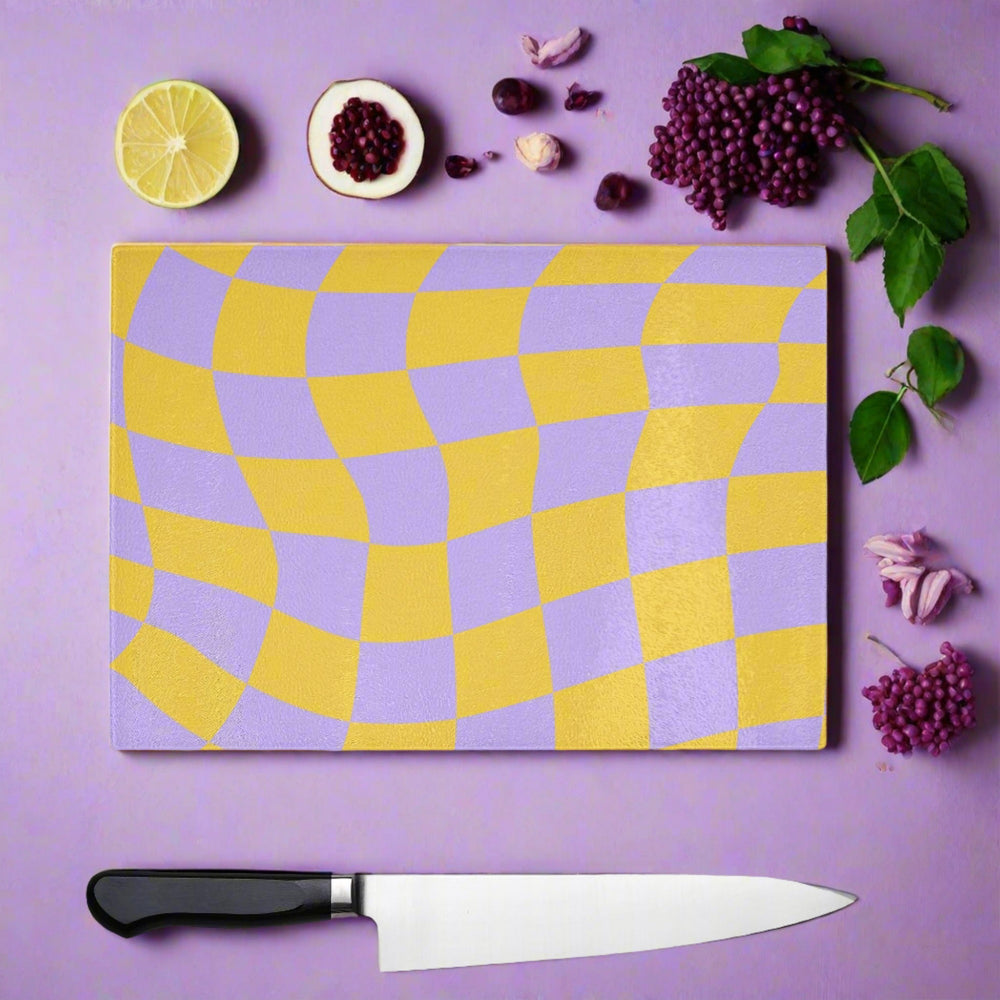 The Yililo Lilac And Yellow Wavy Check Glass Chopping Board adds retro 90s charm to your kitchen, styled on a purple surface with lemon, pomegranate seeds, berries, green leaves, garlic cloves, and a chef’s knife.
