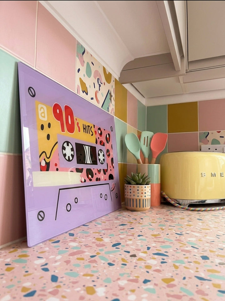 Colourful kitchen with a colourful glass chopping board 90s themed lilac leaned against kitchen tiles with A SMEG toaster in the corner 