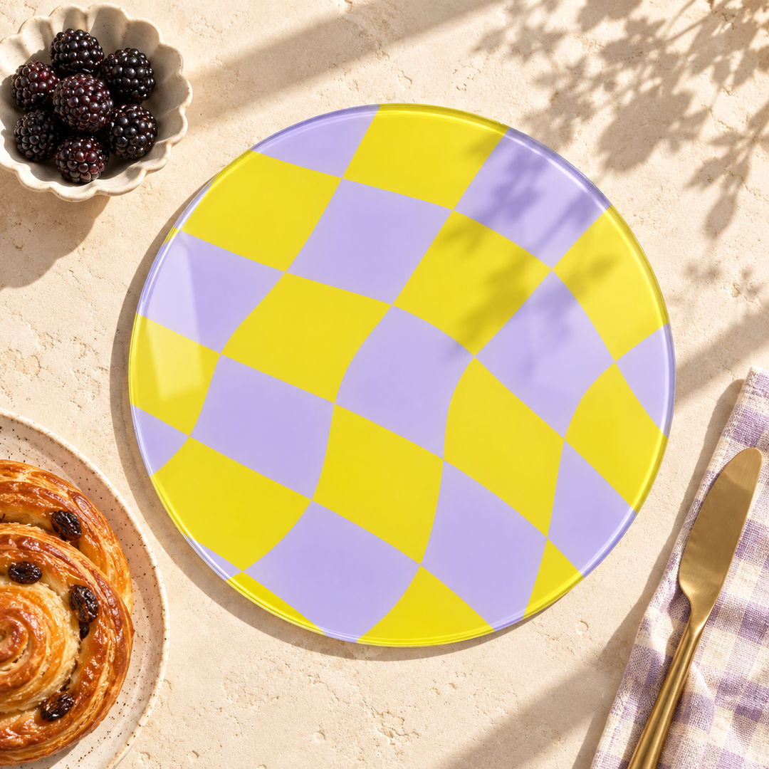 A round glass worktop saver with a yellow and lavender checkerboard pattern, styled on a beige stone surface with a gold knife, a blackberry bowl, and a pastry.