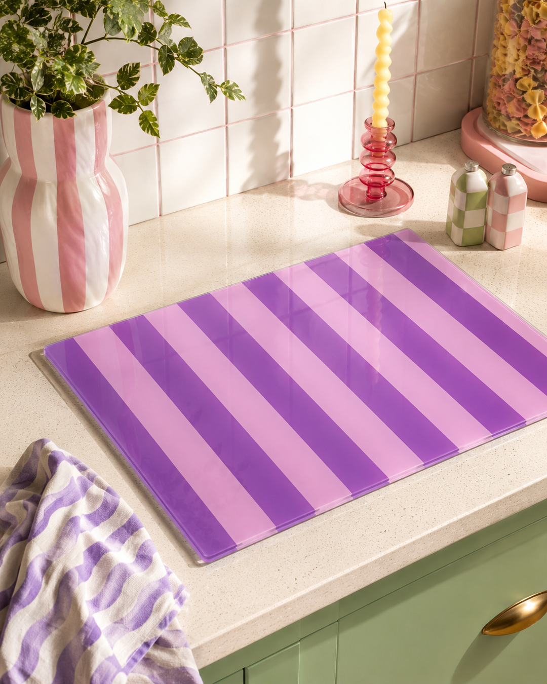 A rectangular glass cutting board with alternating purple and pink diagonal stripes, displayed on a light beige kitchen countertop.