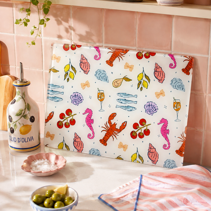 A rectangular kitchen board or tray with a white background and colorful seafood-themed illustration, leaning against a pink tiled kitchen wall. The print includes lobster, seahorse, shell, fish, and cherry motifs.