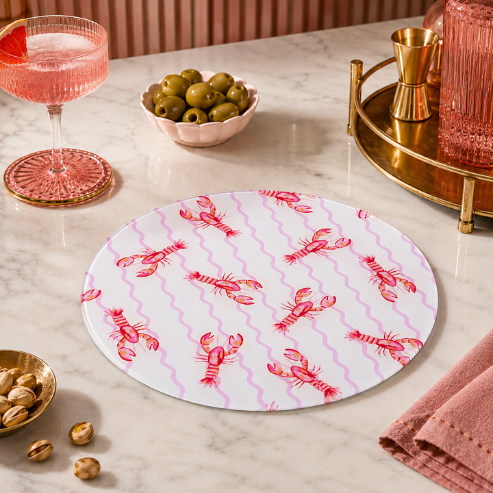 Round glass chopping board with lobster pattern on a marble surface with drinks and snacks.