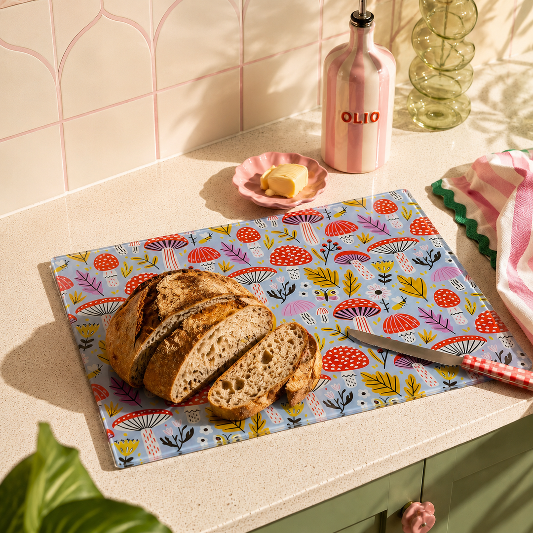 A rectangular chopping board a colourful mushroom and floral pattern sits on a kitchen counter, holding a sliced loaf of bread and a knife. A pink bottle labeled OLIO, butter, and other kitchen items are arranged around it in a styled kitchen setting.