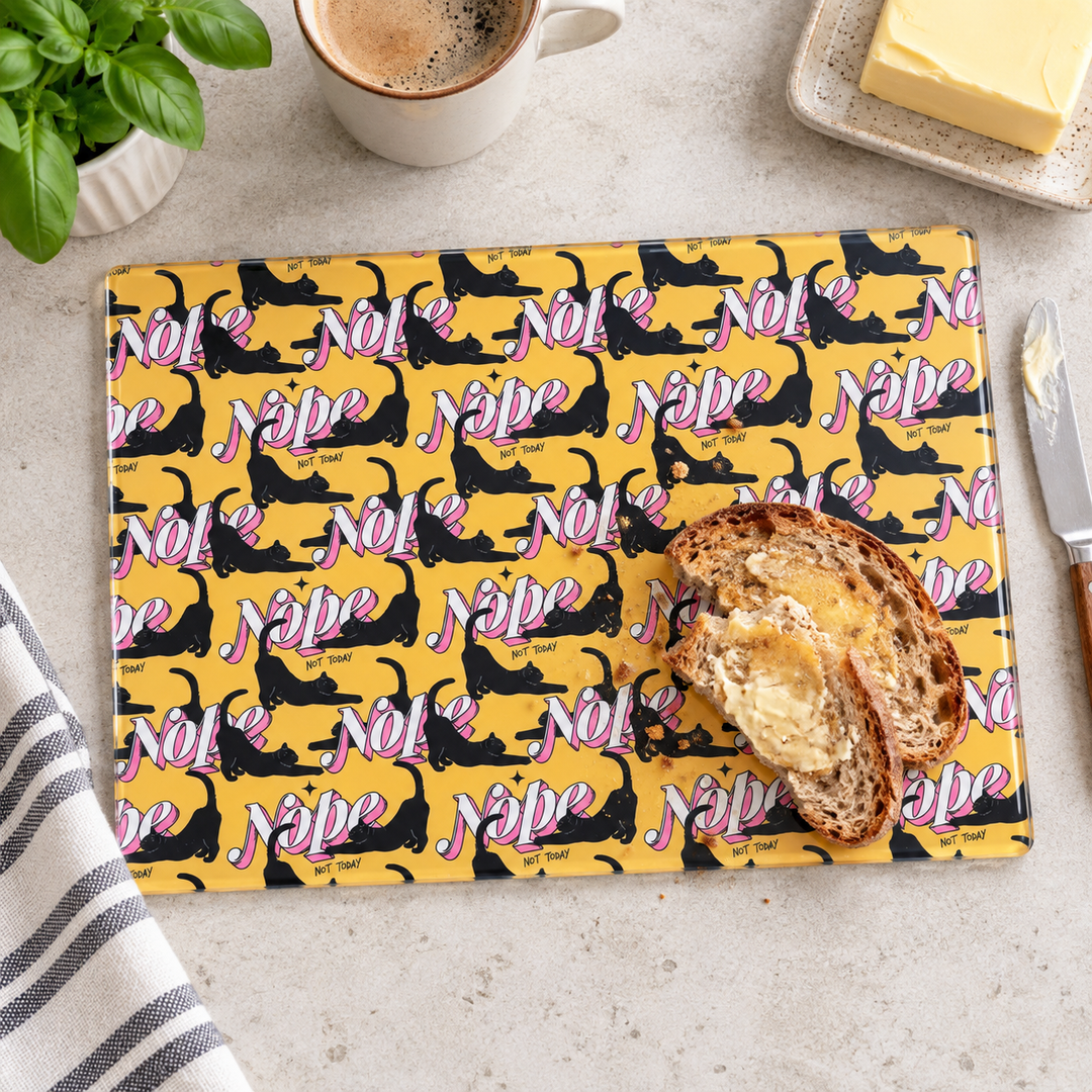 A rectangular glass cutting board with a yellow background, black cat silhouettes, and pink 'Nope' text, styled on a light stone surface with toast, butter, coffee, basil, and a knife.