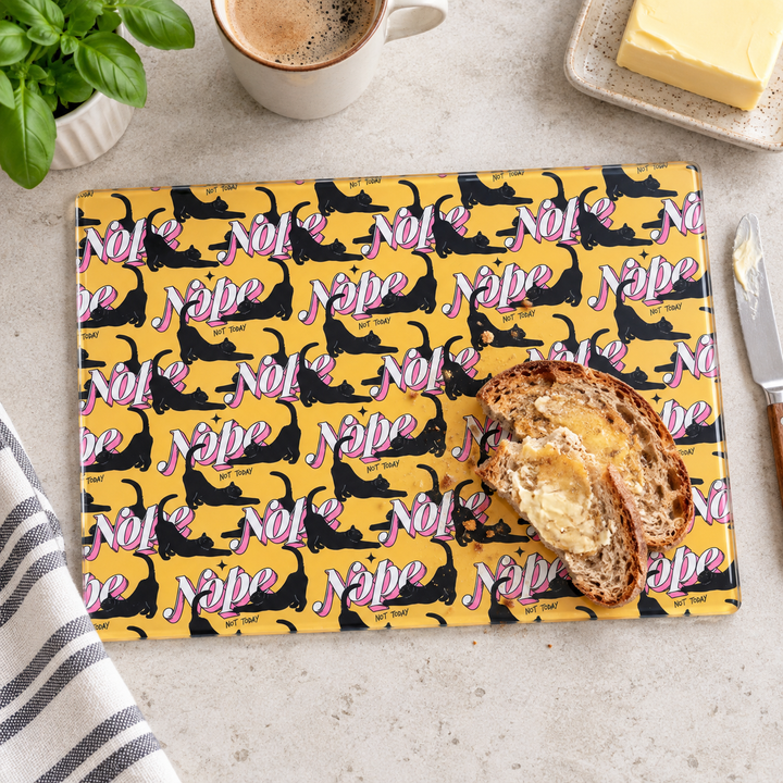 A rectangular glass cutting board with a yellow background, black cat silhouettes, and pink 'Nope' text, styled on a light stone surface with toast, butter, coffee, basil, and a knife.