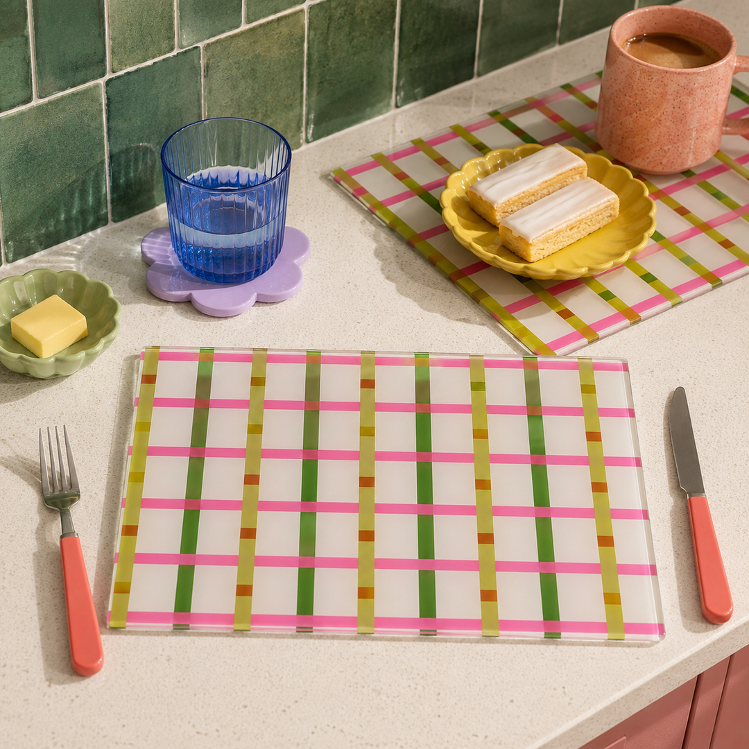 A glass placemat set with a pink, green, and gold striped grid pattern, styled on a kitchen counter with a fork, knife, glass, and food.