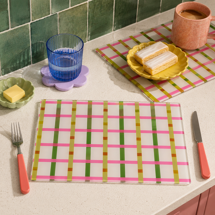 A glass placemat set with a pink, green, and gold striped grid pattern, styled on a kitchen counter with a fork, knife, glass, and food.