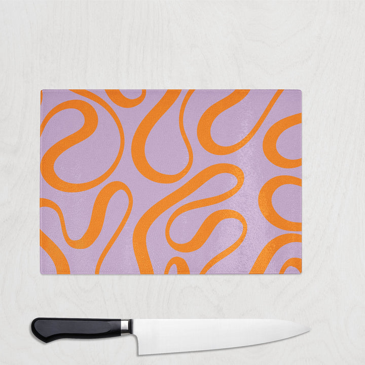 The Yililo Orange Loop Y2K Glass Chopping Board, featuring an orange swirly pattern on lavender, rests on a light surface. Below it lies a heat-resistant chef’s knife with a black handle.