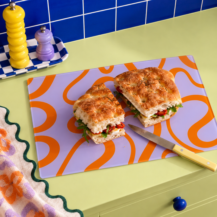 The Yililo Orange Loop Y2K Glass Chopping Board, featuring an orange swirly pattern on lavender on a lime green work surface with a sandwich on top with a knife and a tea towel with a scallop edge beside it. heat-resistant worktop saver