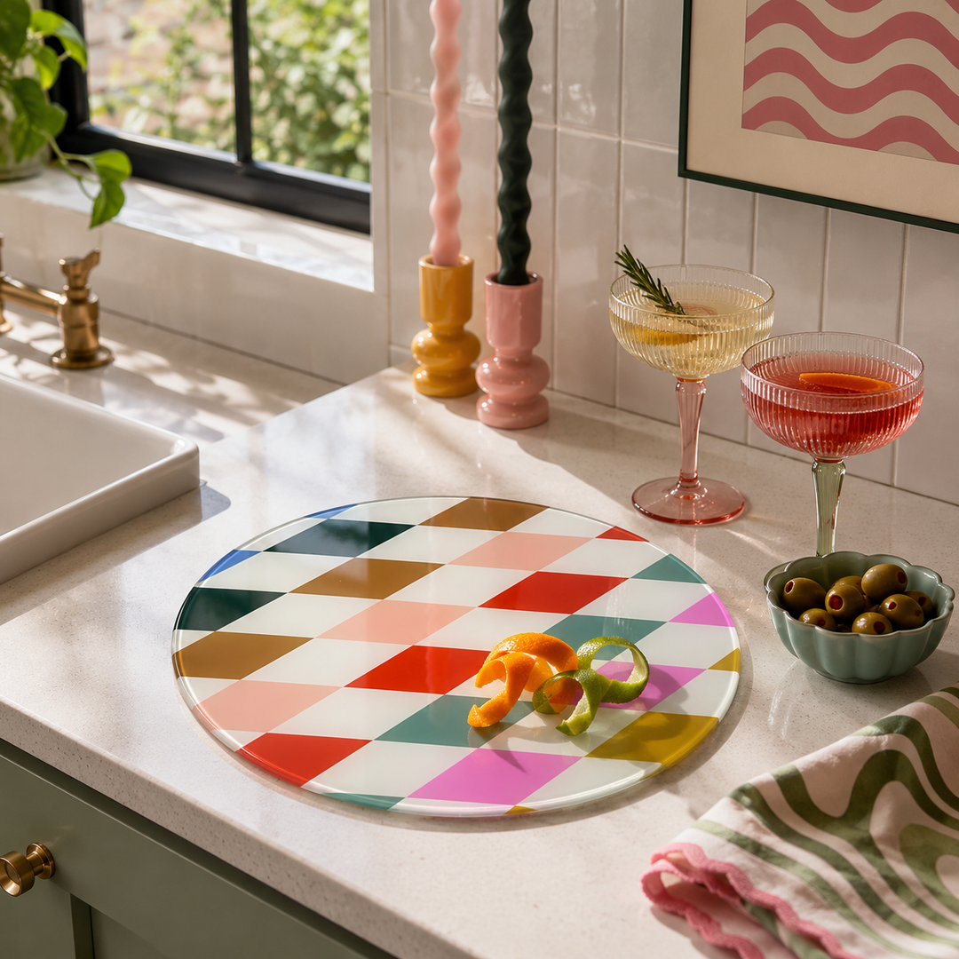 A round glass serving platter glass chopping board with a colourful geometric diamond pattern sits on a kitchen counter next to two filled coupe glasses, a bowl of olives, and a striped cloth.
