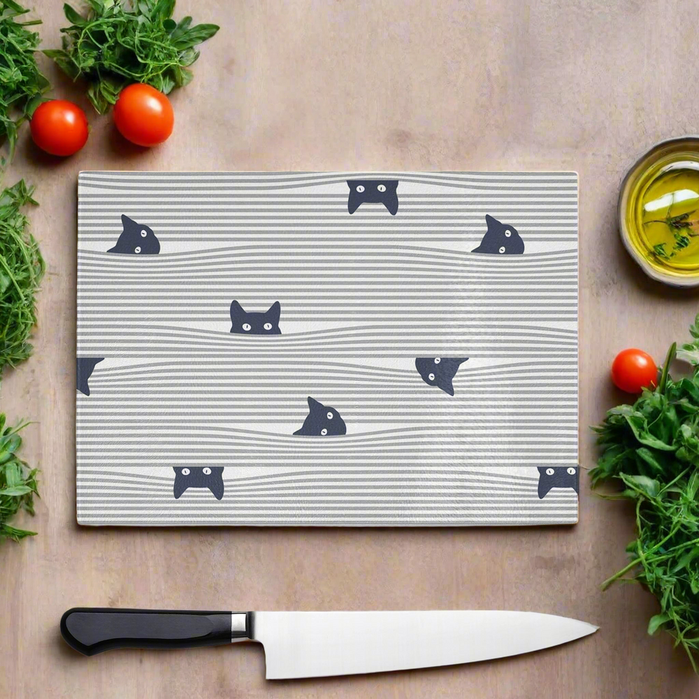 The Yililo Peeping Black Cat Glass Chopping Board features adorable cat faces behind wavy lines, ideal for cat lovers. Shown with herbs, tomatoes, olive oil, and a chef’s knife on a wooden surface.