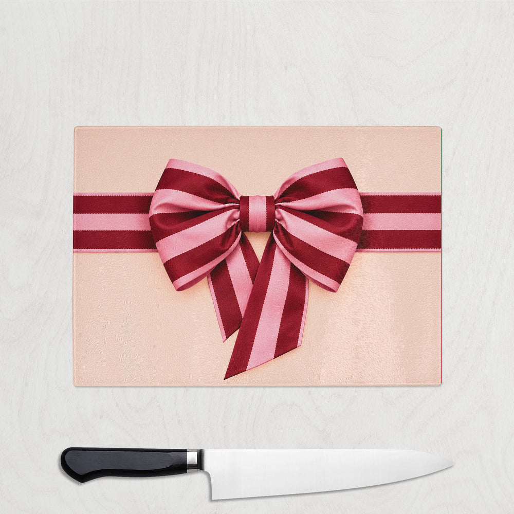 Pink And Burgundy Bow Christmas glass chopping board with a pink and burgundy striped satin ribbon and bow on a blush pink background, shown with a chef's knife