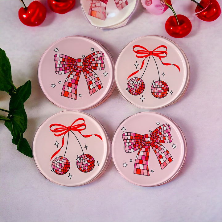 The Yililo Pink Disco Cherry And Bow 4 Piece Glass Coaster Set features vibrant pink round coasters with cherry, bow, ribbon, and disco ball designs, shown on a white surface with artificial cherries and green leaves.
