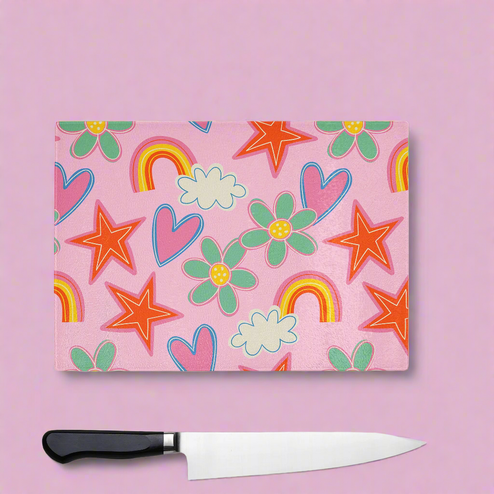 The Yililo Pink Doodle Glass Chopping Board Worktop Saver, featuring red stars, green and yellow flowers, pink hearts, rainbows, and clouds on a pink background, adds a quirky touch to your kitchen. A large black-handled knife lies below it.