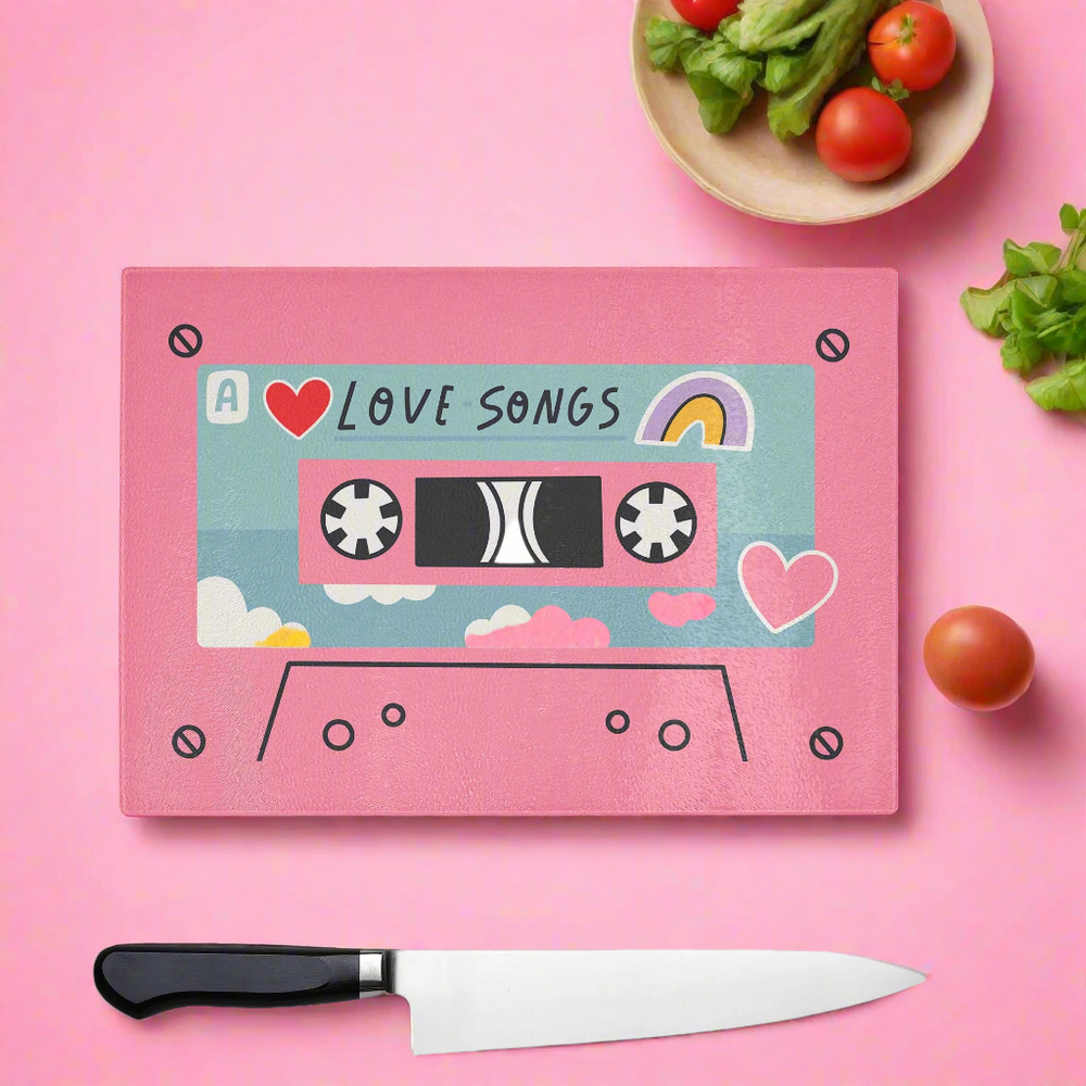 The Yililo Pink Love Songs Cassette Tape Glass Chopping Board features a quirky retro cassette design with hearts and a rainbow, perfect for 90s music fans. Shown with veggies, a plate, and knife on a pink background.