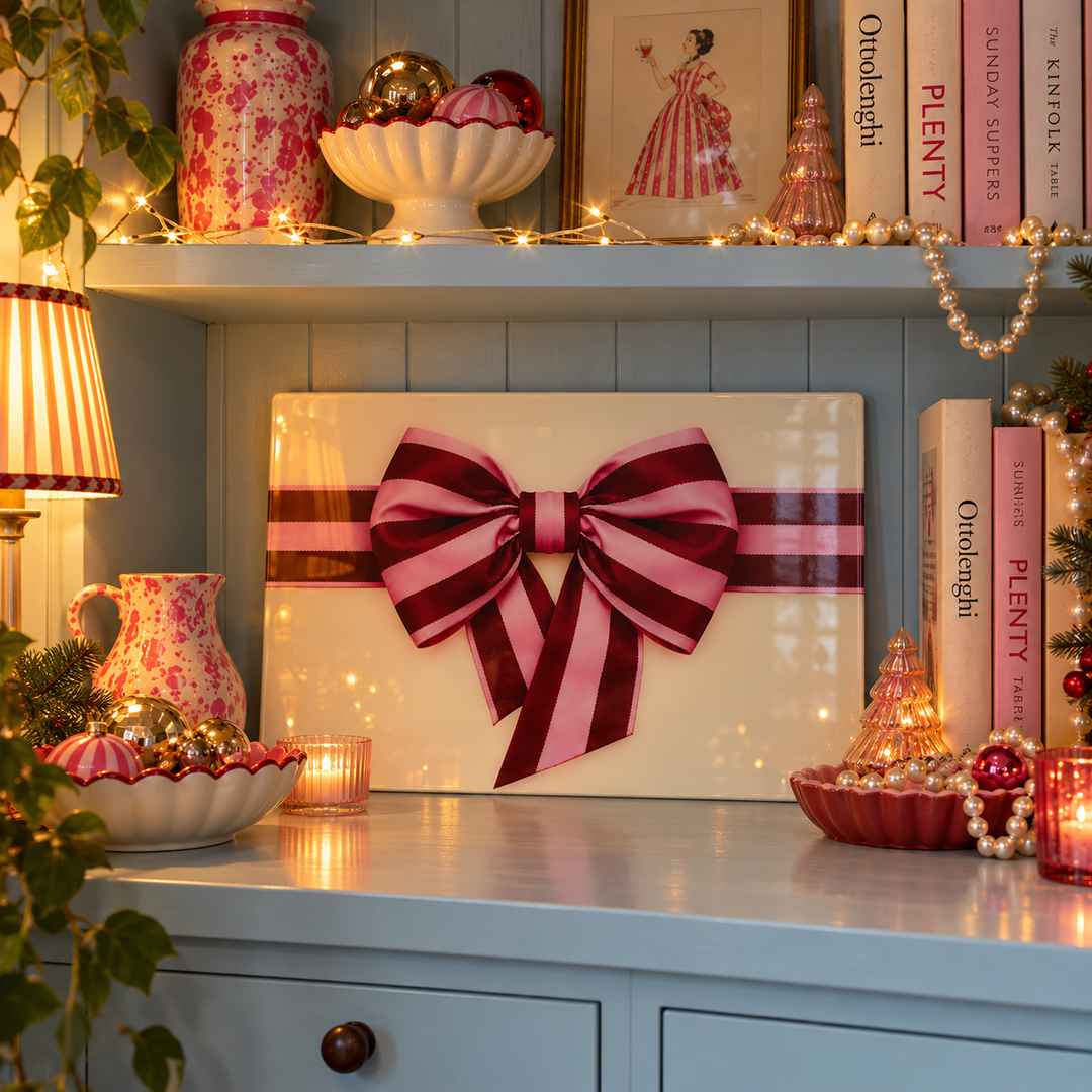 A square cream-colored decorative panel or sign with a large pink and burgundy striped bow across the center, displayed on a blue cabinet in a decorated holiday room with candles, books, and ornaments.