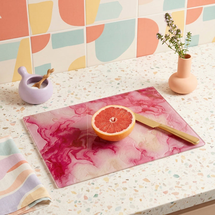 Glass cutting board with pink and cream abstract design on a speckled countertop with a half-sliced grapefruit and knife