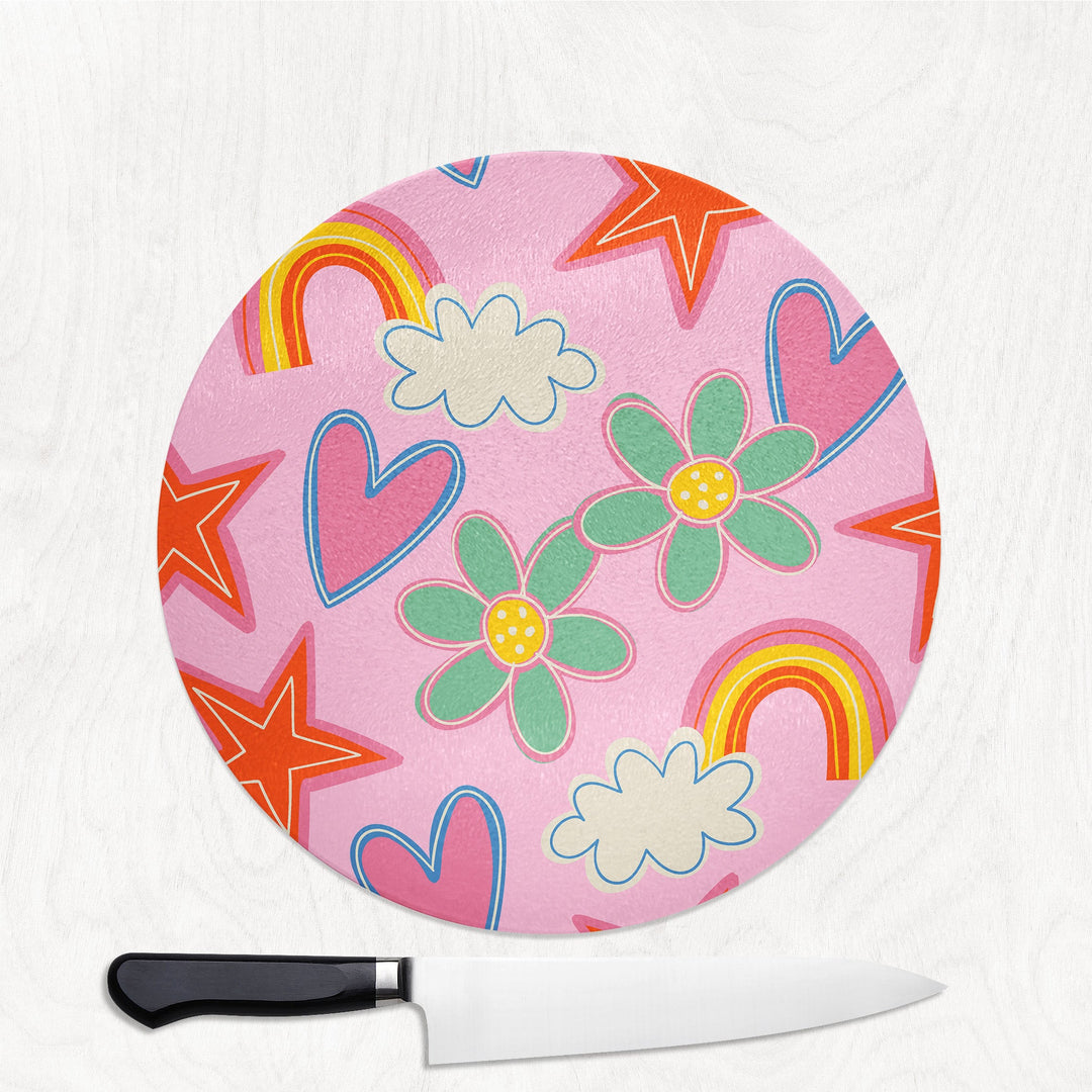Round pink cutting board with colorful floral and star designs, accompanied by a knife on a white background.