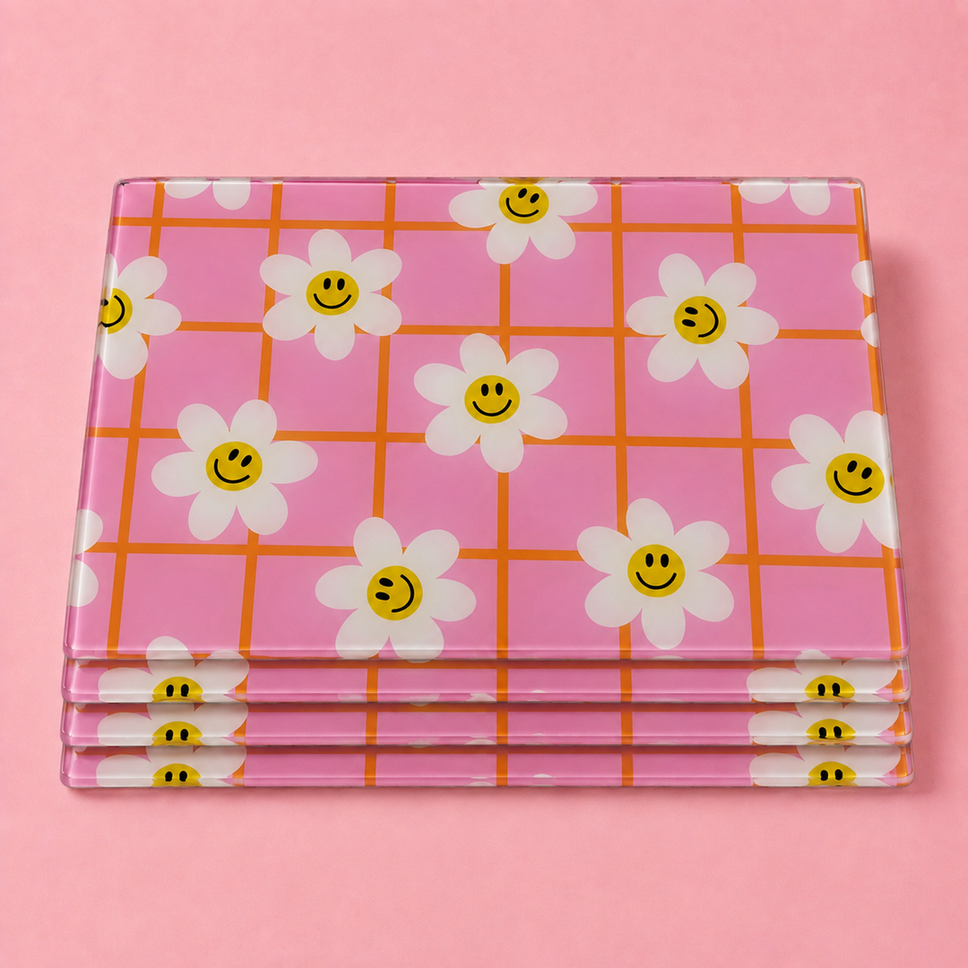 A set stack of rectangular glass pink tablemats with a white daisy and orange grid pattern, photographed on a solid pink background.
