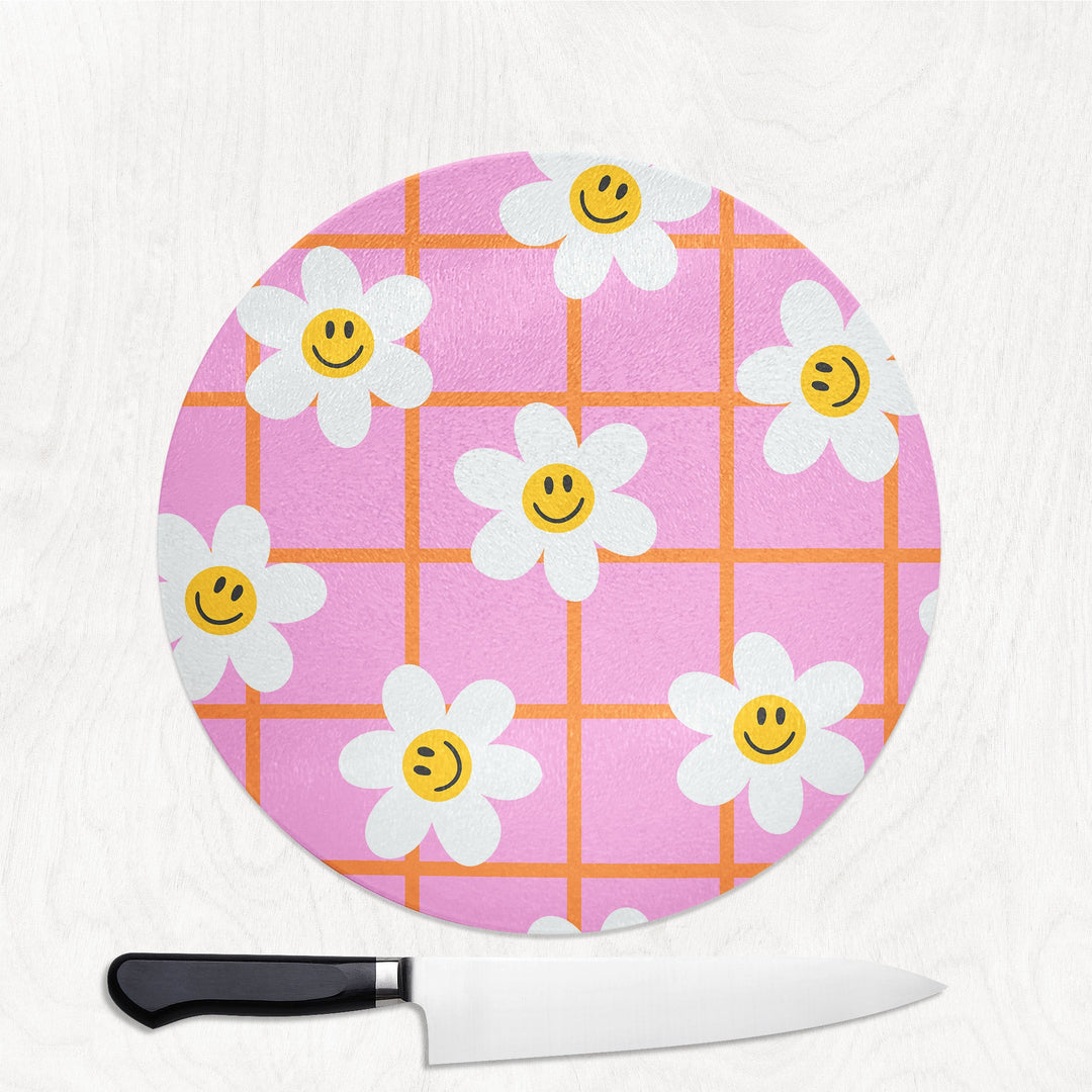 Round cutting board with smiley face flowers on a pink and orange checkered pattern, accompanied by a knife.