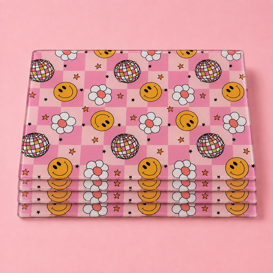 A stack of rectangular glass tablemat set with a pink checkerboard pattern featuring yellow happy faces, white daisies, disco balls, and small stars, displayed on a solid pink background.