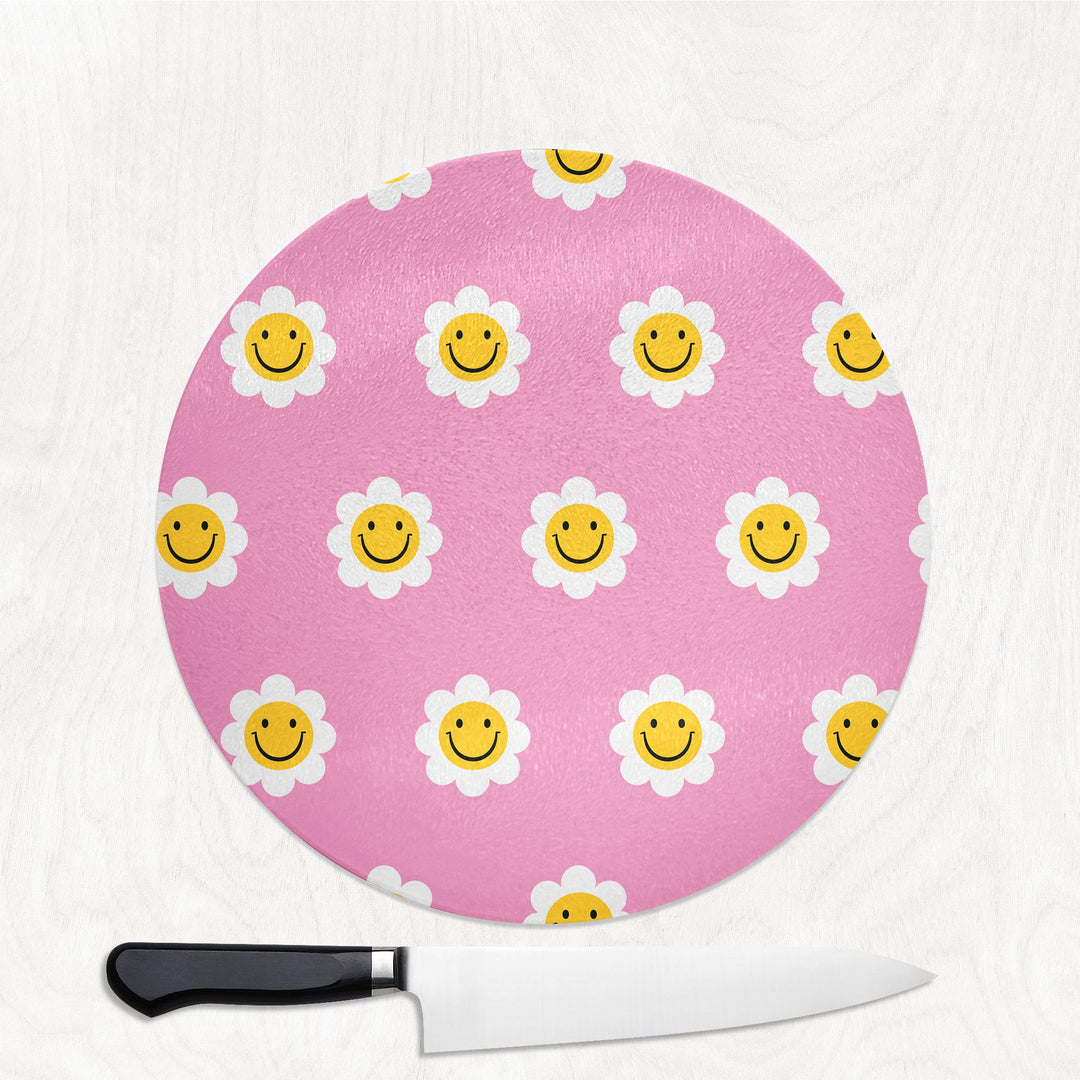 Pink round cutting board with smiley face daisy pattern and a knife on a white background