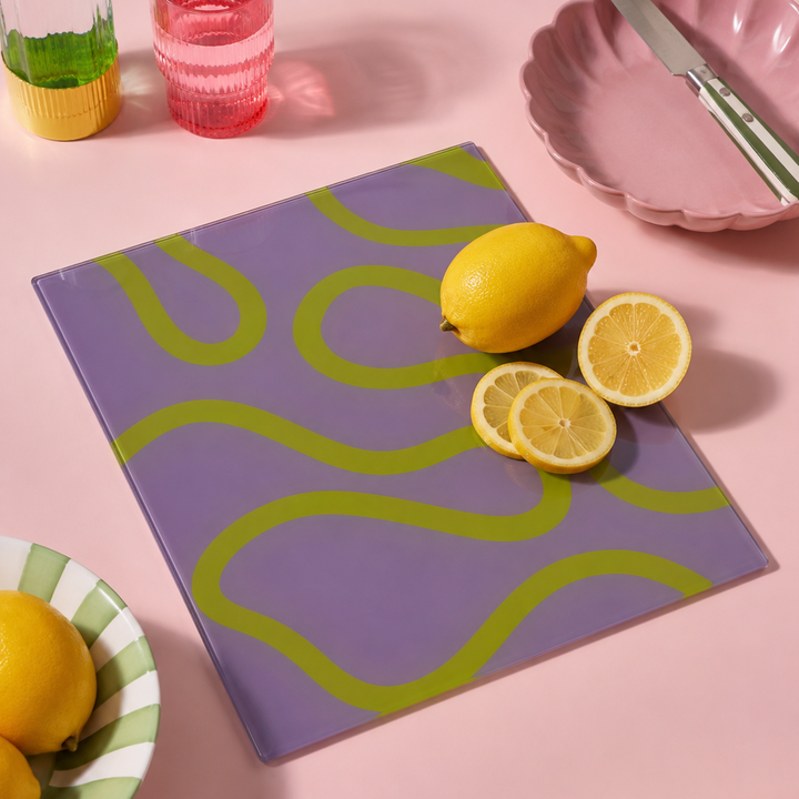 Purple cutting board with green wavy lines on a pink surface with lemons and a glass.