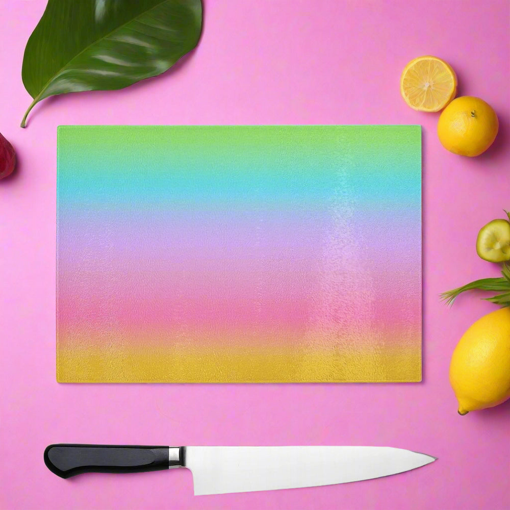 The Yililo Rainbow Bliss Glass Chopping Board adds vibrant color to a pink surface, surrounded by a chef’s knife, lemon halves, cucumber, leafy greens, and a large green leaf.