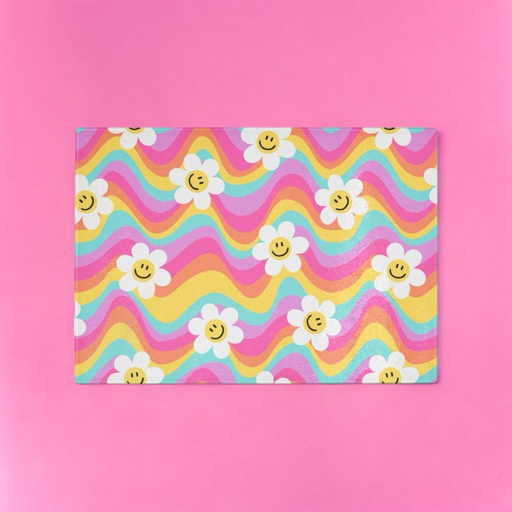 The Yililo Rainbow Wave Flower Smiley Glass Chopping Board features a wavy pastel pattern with smiling daisies on a solid pink background, adding a cheerful touch to any colourful kitchen.