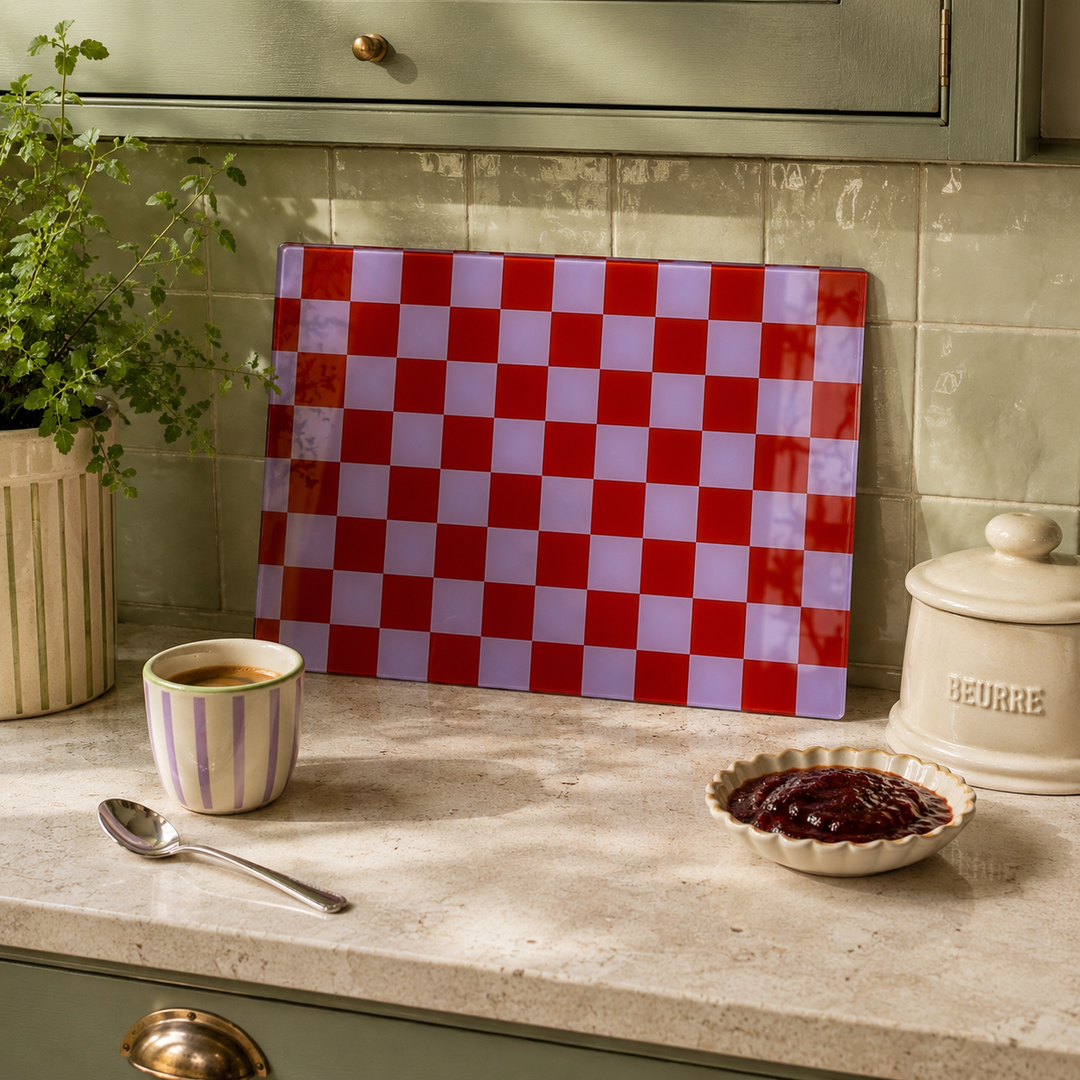 A glass cutting board with a red and lavender checkerboard pattern, displayed in a styled kitchen setting on a marble countertop next to a striped mug, a spoon, and a jar of jam.
