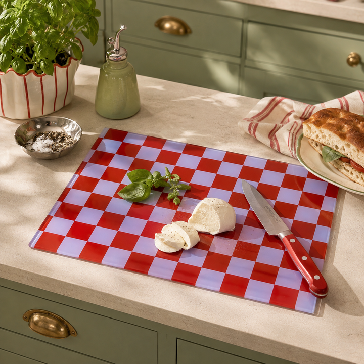 A red and lavender checkered glass cutting board on a beige countertop, styled with a knife, fresh mozzarella, basil, a potted plant, and a sandwich in a green kitchen setting.