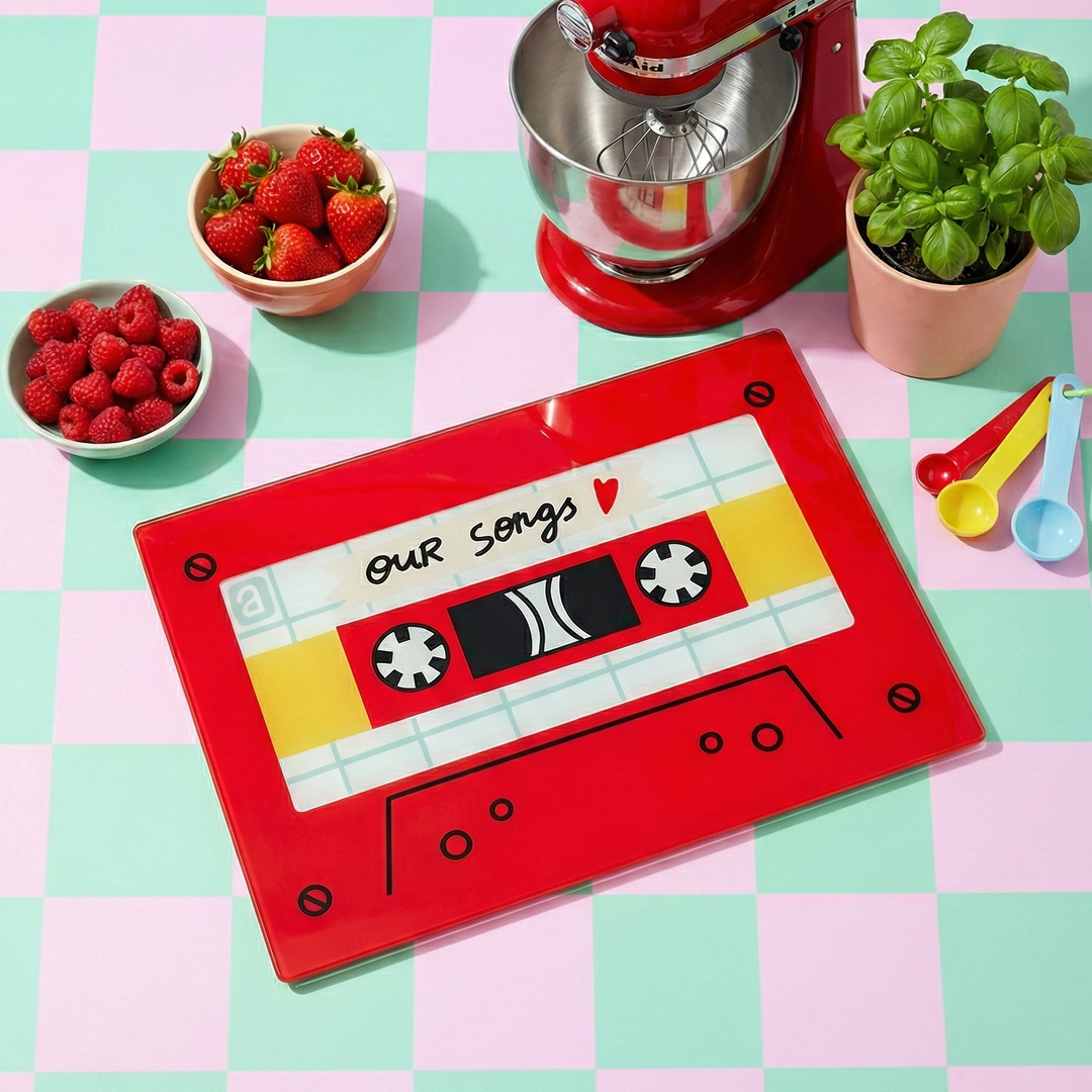 Red cassette tape-shaped placemat with 'Our Songs' text on a checkered tablecloth with kitchen items.