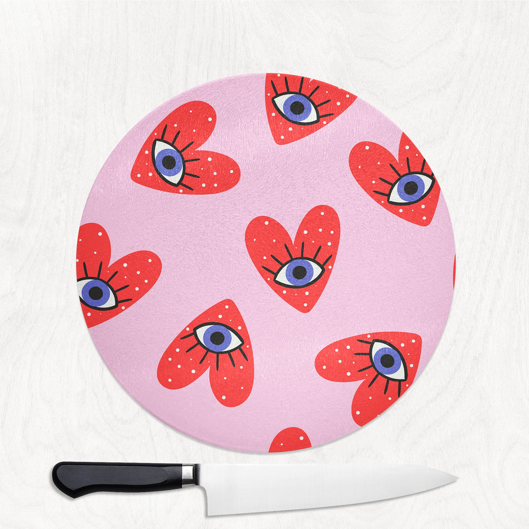 Pink cutting board with red heart designs and blue eyes on a white background, accompanied by a knife.