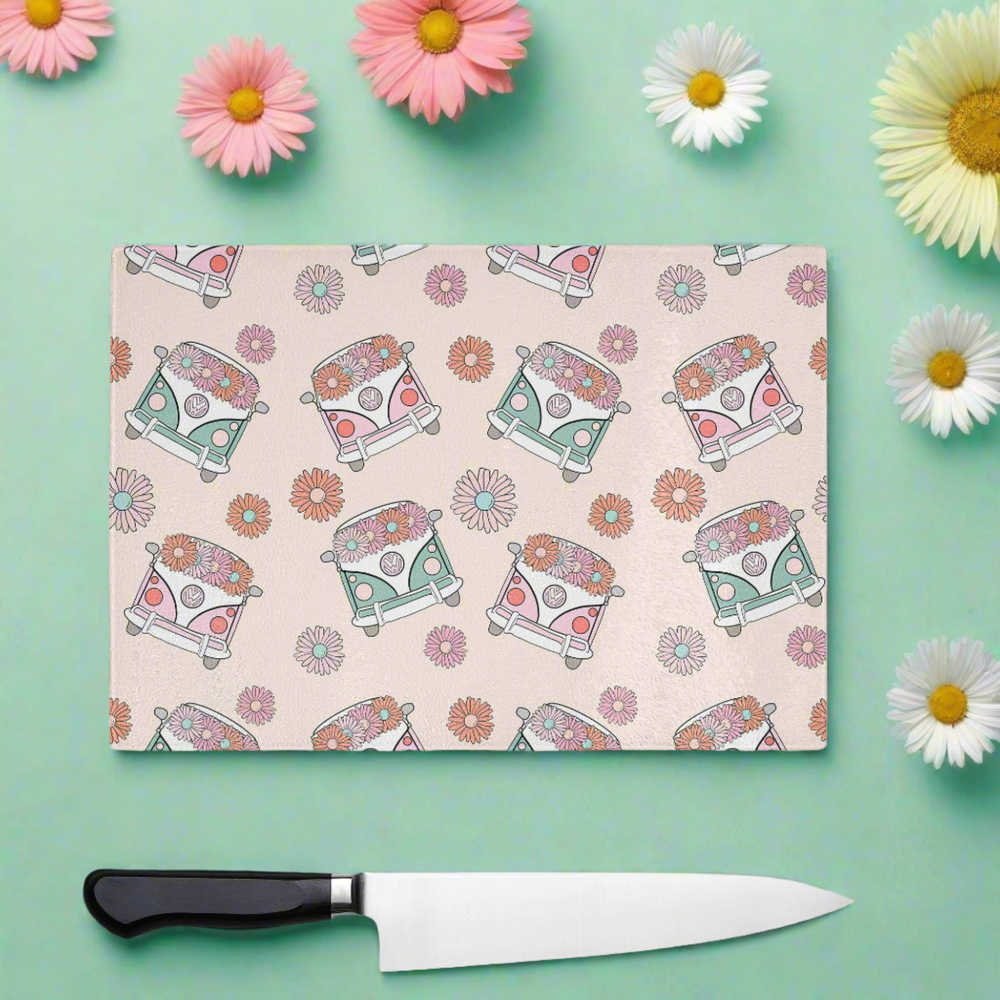 The Yililo Retro Camper Van Glass Chopping Board, featuring colorful vans and daisies, sits on a mint green surface with real daisies nearby. Ideal for VW fans and retro kitchens, a knife is placed just below the board.