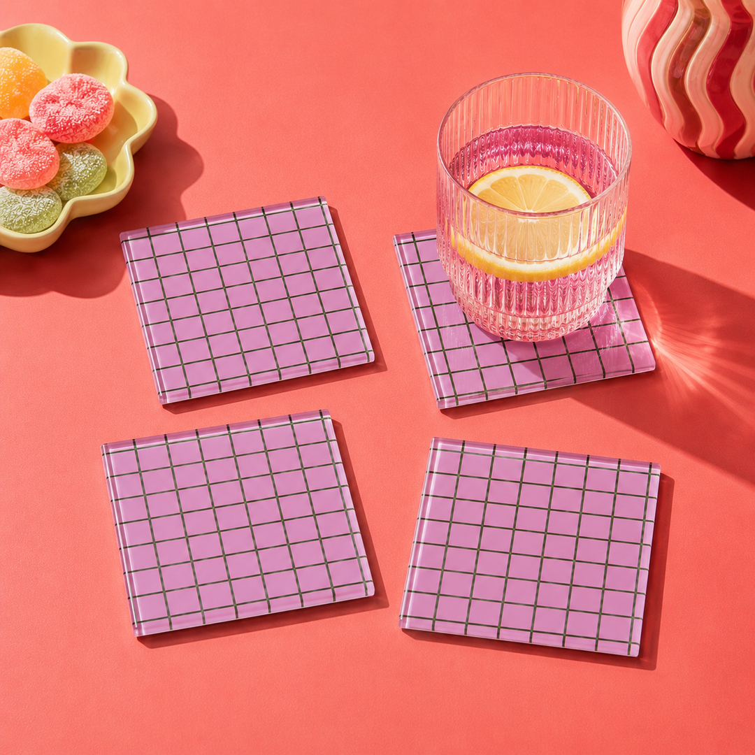 Four square pink coasters with a green grid pattern arranged on a coral background, with a glass of pink drink and lemon on one coaster. A yellow flower-shaped candy dish and a striped object appear at the edges as styled props.