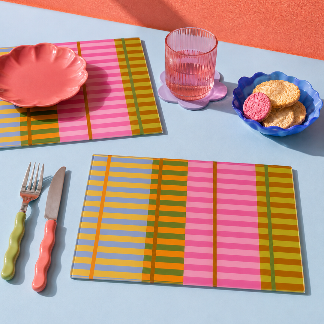 Colourful glass striped placemats and matching cutlery arranged on a light blue table with a pink wall background. The set includes a scalloped pink plate, a ribbed pink glass tumbler on a flower-shaped coaster, cookies in a blue dish, and utensils with pink, green, and orange handles.