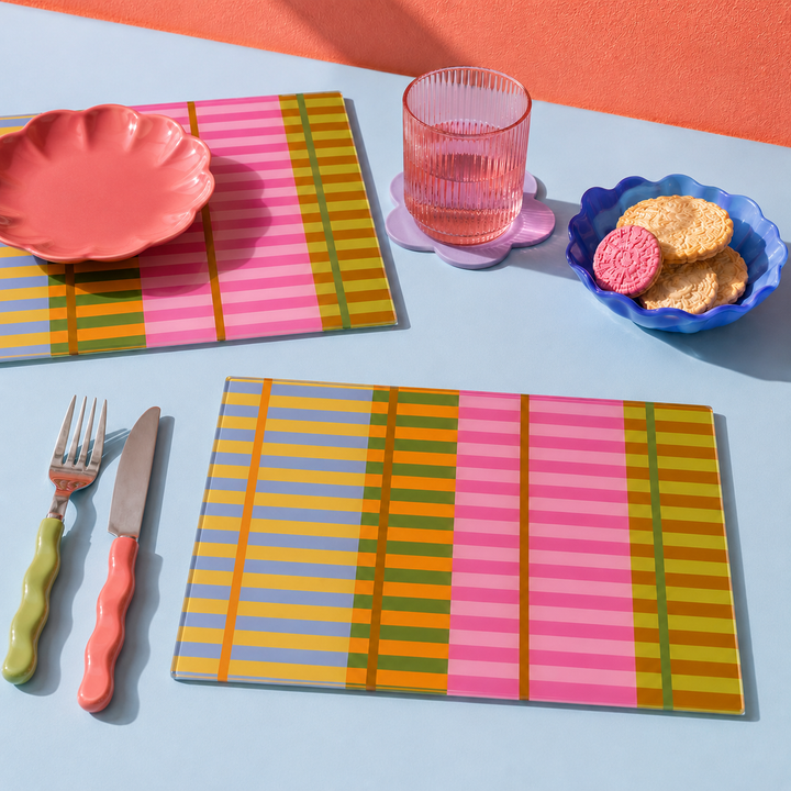 Colourful glass striped placemats and matching cutlery arranged on a light blue table with a pink wall background. The set includes a scalloped pink plate, a ribbed pink glass tumbler on a flower-shaped coaster, cookies in a blue dish, and utensils with pink, green, and orange handles.
