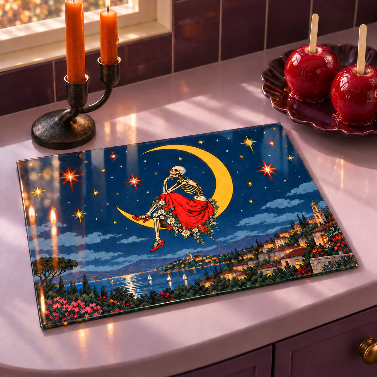 A rectangular glass cutting board with a colorful Halloween design of a skeleton riding a crescent moon over a lake, displayed on a kitchen counter next to candles and apple cider apples.