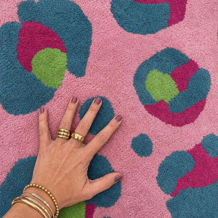 Hand with gold rings and bracelets on a pink rug with blue and green leopard print patterns