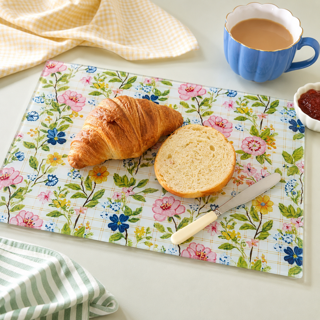 A rectangular glass cutting board with a colourful floral print, styled with a croissant, a slice of bread, a knife, a blue cup of coffee, and a small dish of jam on a light tabletop by Yililo.