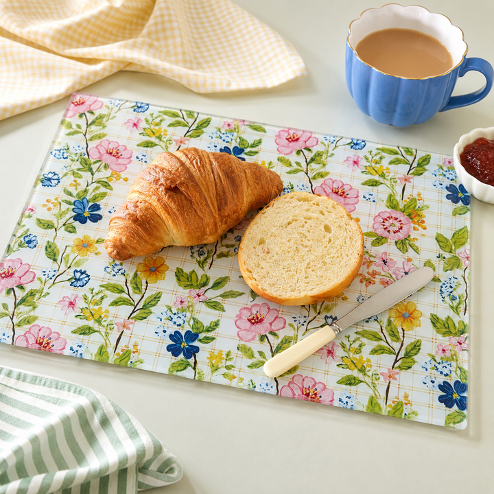 A rectangular glass cutting board with a colourful floral print, styled with a croissant, a slice of bread, a knife, a blue cup of coffee, and a small dish of jam on a light tabletop by Yililo.
