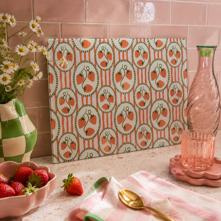 A rectangular chopping board with a repeating strawberry pattern, standing on a countertop in a styled kitchen-like setting. The board has a white background with vertical coral-red stripes and oval motifs containing strawberries with green stems and leaves.
